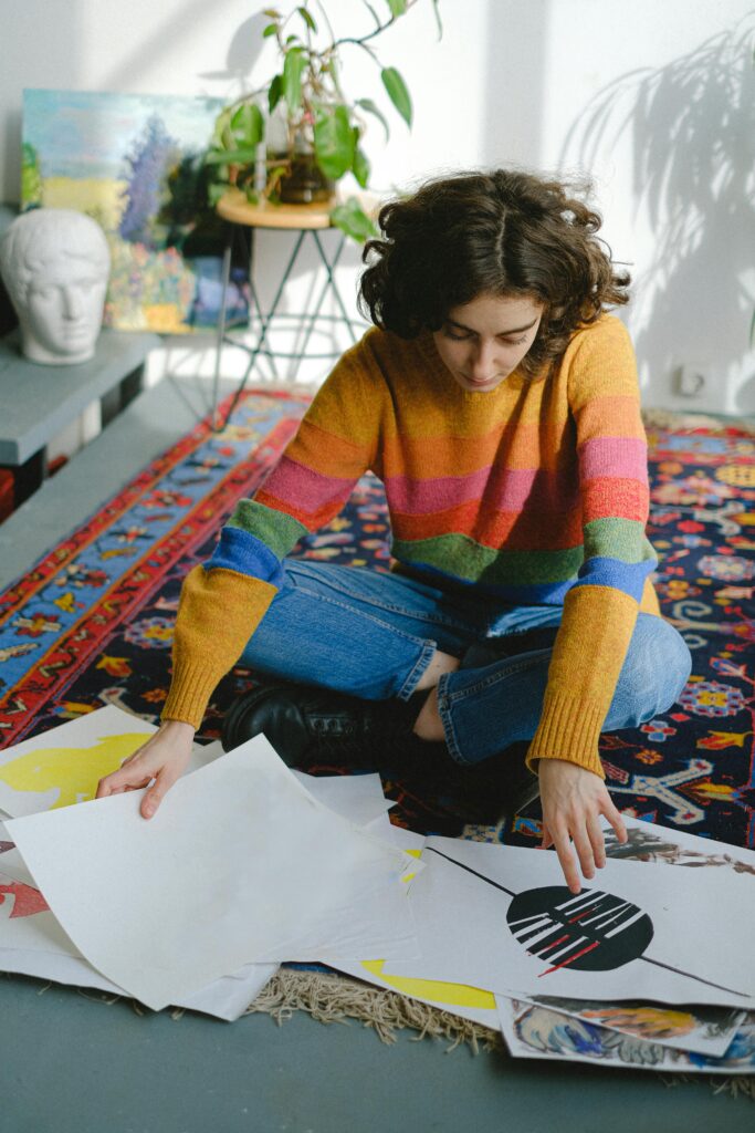 High angle full length of young thoughtful woman in casual clothes sitting on carpet on floor while looking through paintings near head sculpture and potted plant in daylight in room